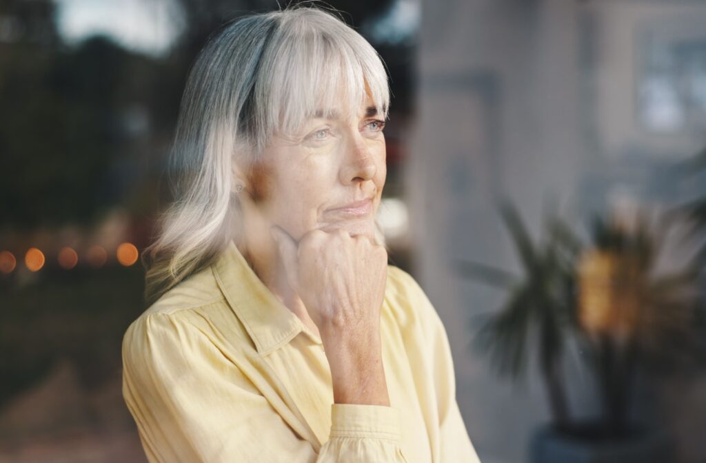 Person with grey hair looking out a window thoughtfully with hand on chin