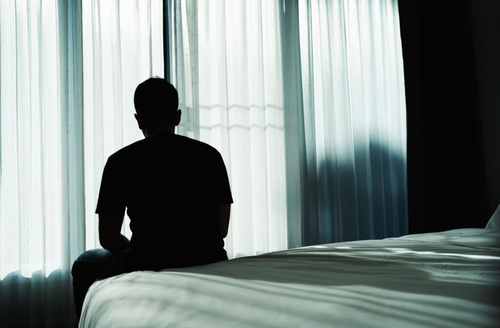 Silhouetted person sitting on edge of bed staring out sheer-curtained window in a dark contemplative mood