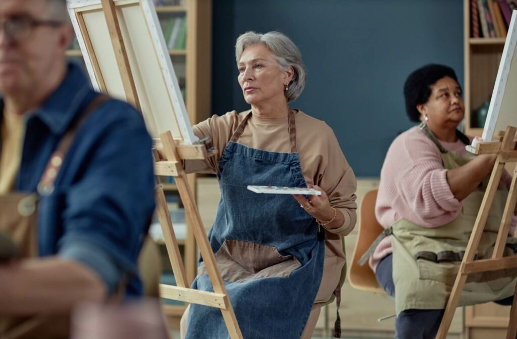A senior adult paints on an easel in an art class with a group of seniors enjoying a social activity in their personal care community.