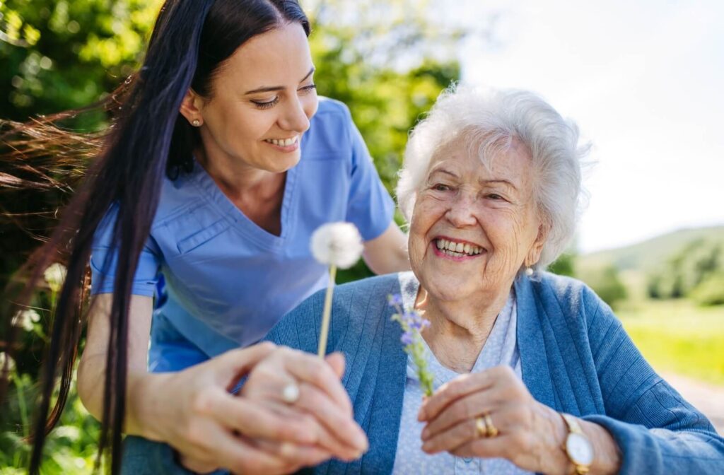 A caregiver hands a smiling older adult a dandelion while taking a walk together in the outdoor grounds of an assisted living community