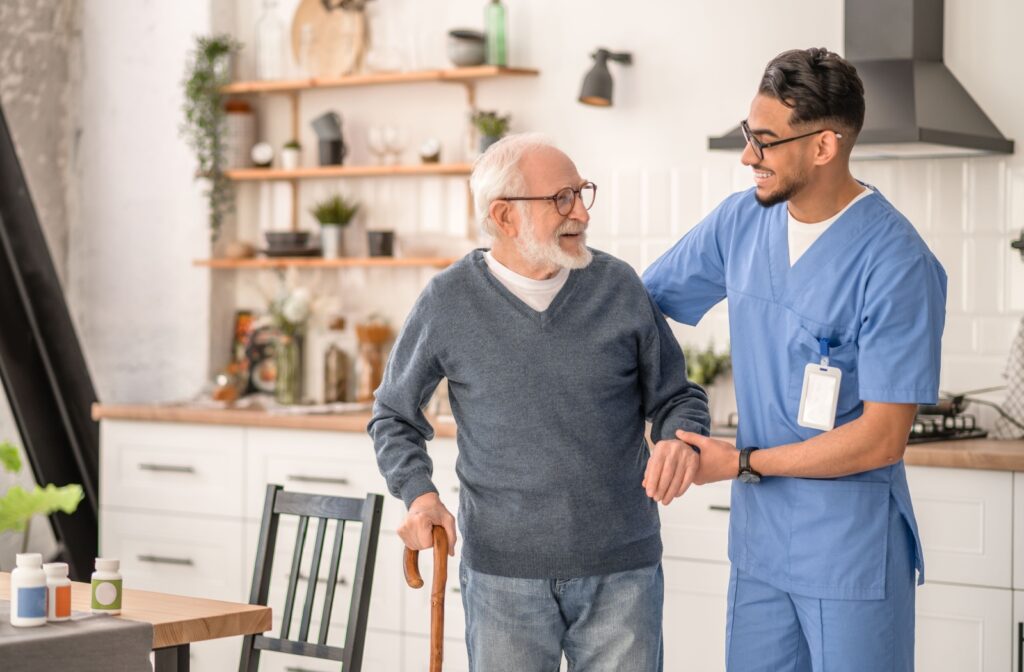 A caregiver helps a smiling resident balance their cane in their kitchen in their home in assisted living