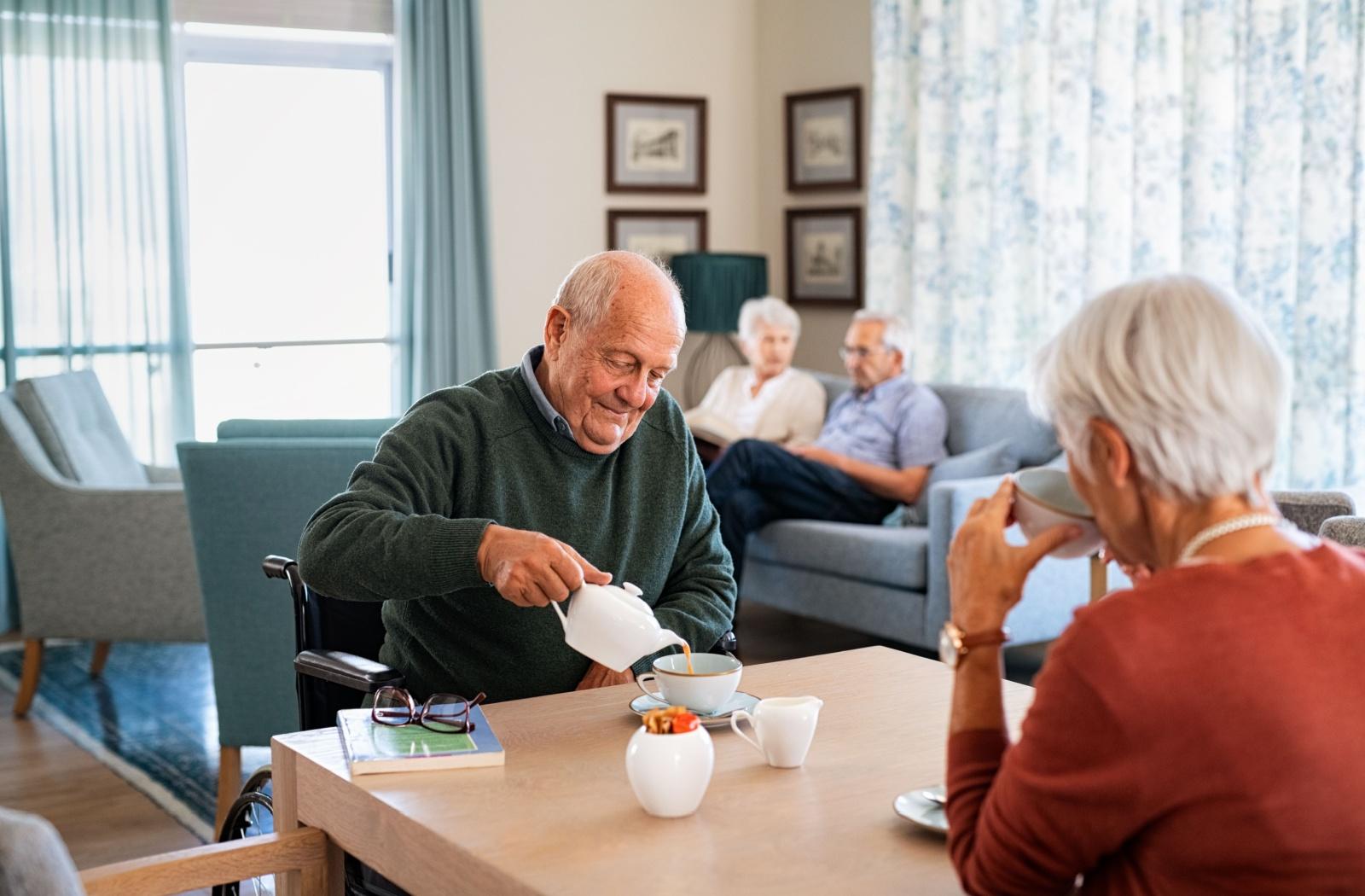 A senior pours a cup of tea while sitting at a table with their spouse.
