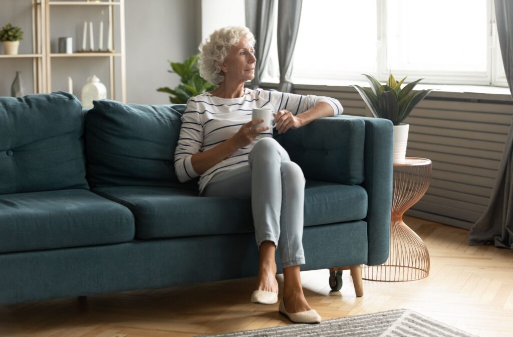 A senior adult looks wistfully out of a window while sitting on a dark teal sofa.